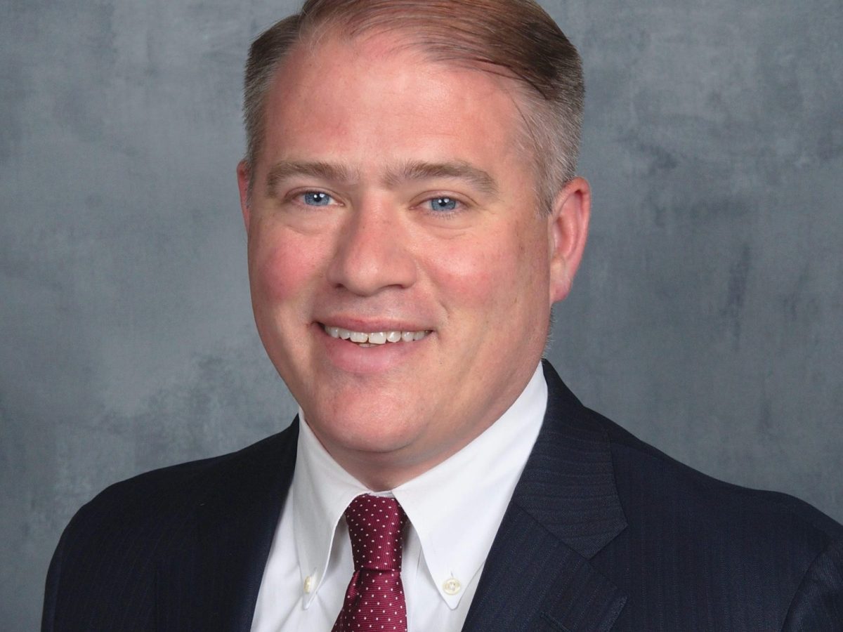 headshot of a man smiling while wearing a suit and tie against a gray background.