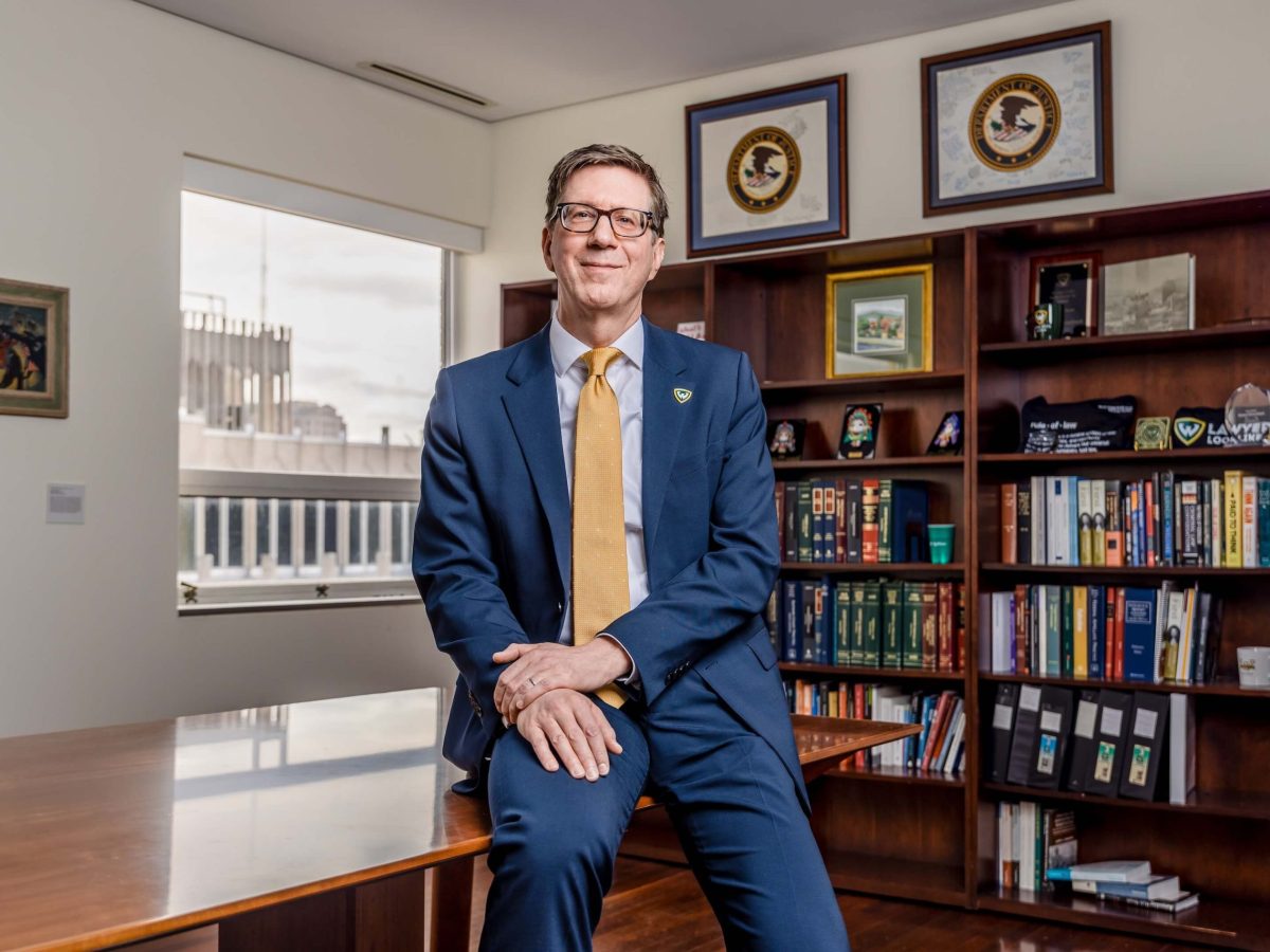 A smiling man sits on a desk in front of a window and a bookcase