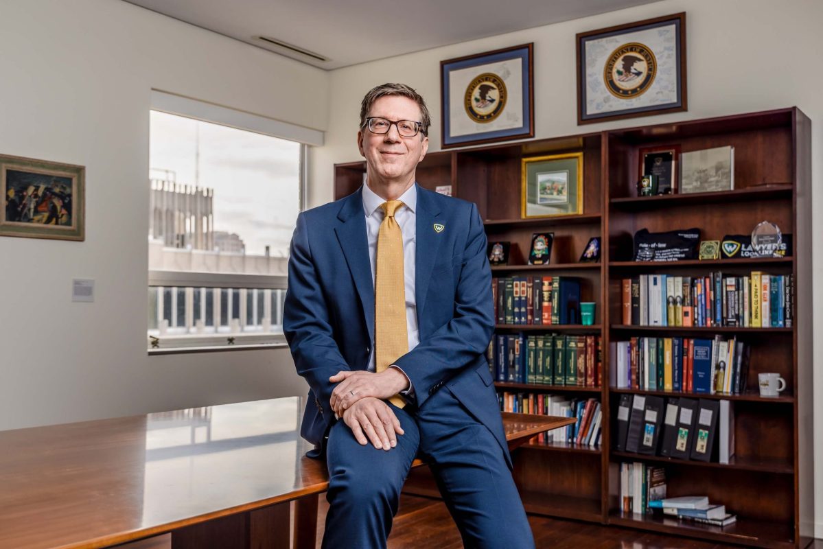 A smiling man sits on a desk in front of a window and a bookcase
