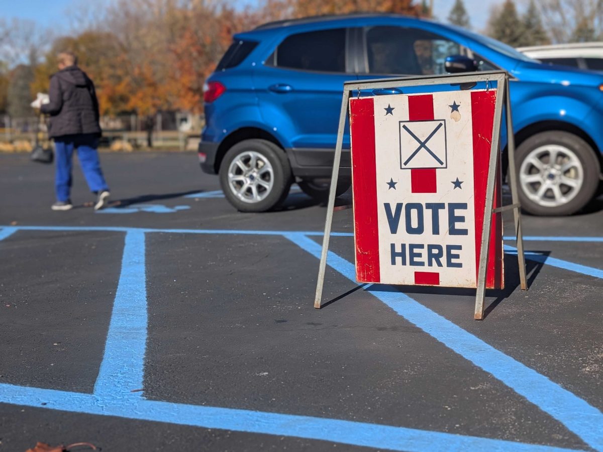 A Vote Here sign in a parking lot. In the background is a blue car and a voter who appears to be walking away.