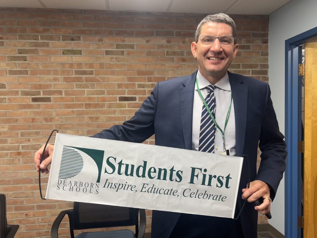Smiling man holding up a Dearborn Public Schools reading "Students First Inspire, Educate, Celebrate"