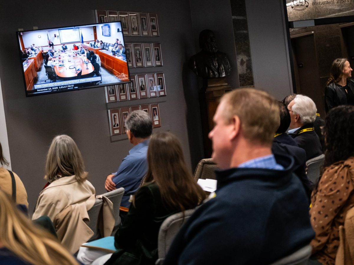 People sit in an overflow room watching a monitor showing a meeting occurring in an adjacent room.