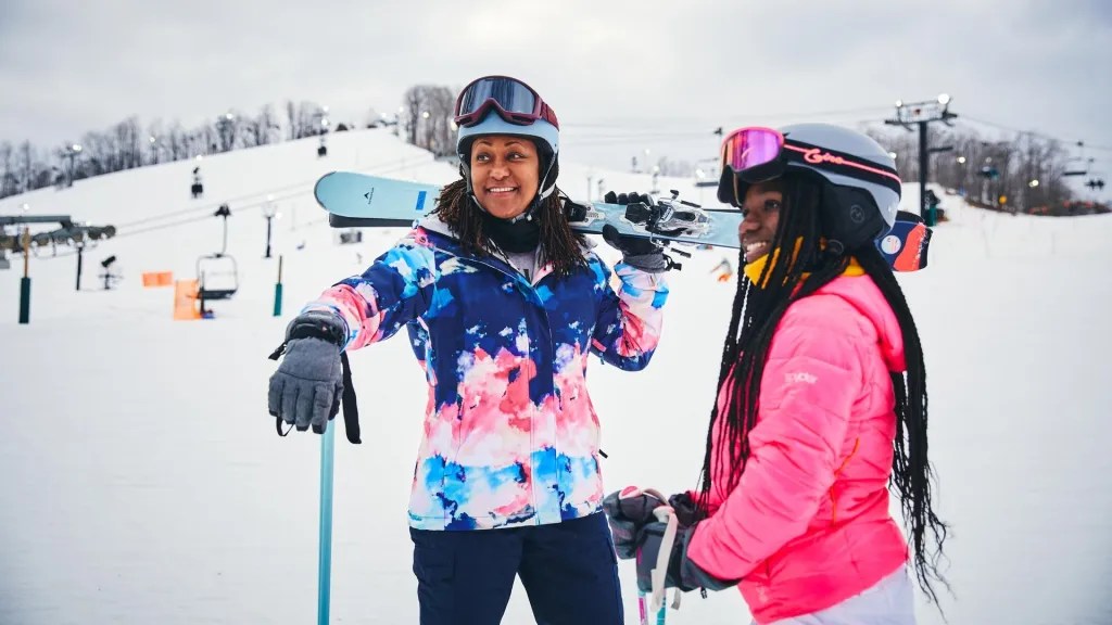 Two people stand at the bottom of a ski hill in winter gear