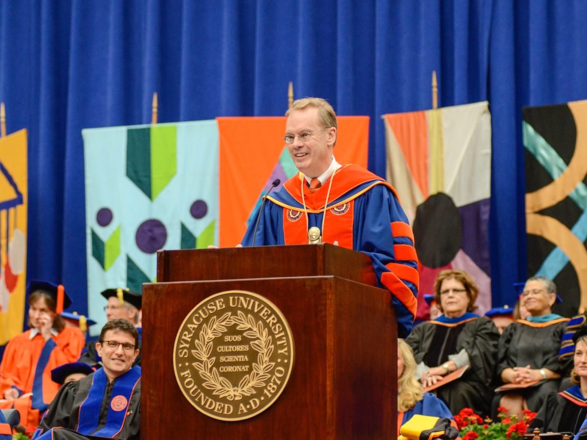 A man dressed in commencement regalia stands at a podium with a Syracuse University crest affixed to the front.