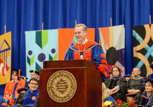 A man dressed in commencement regalia stands at a podium with a Syracuse University crest affixed to the front.