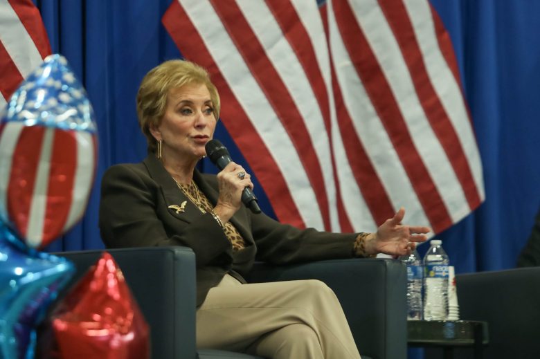US Education Secretary Linda McMahon sits on stage and speaks into a microphone while surrounded by an American flag and red, white and blue balloons.