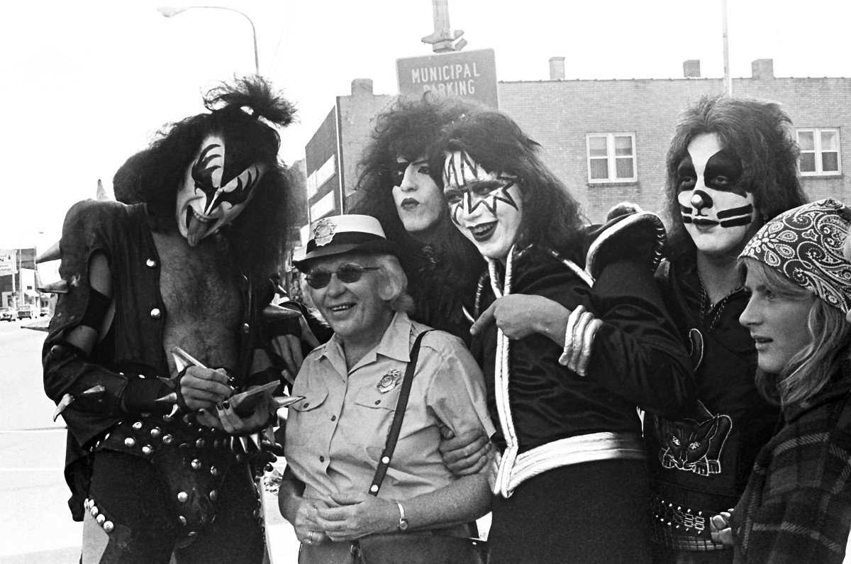 A black and white photo shows the band KISS posing for a photo with a Cadillac resident