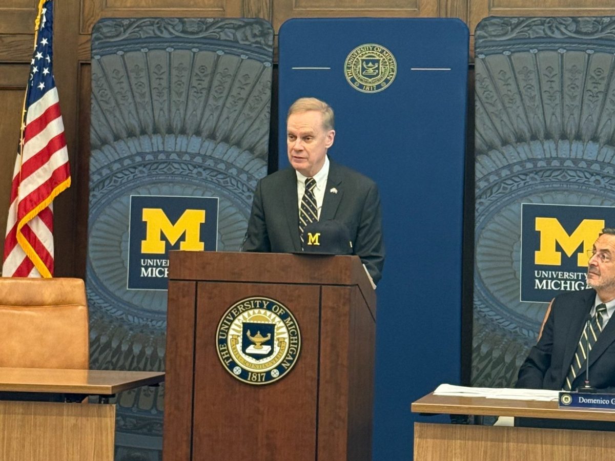 A man at a podium surrounded by University of Michigan signage and mementos.