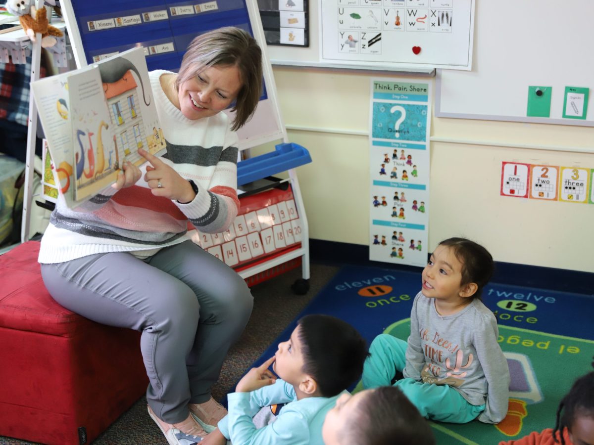 A teacher points to an illustration in a book as pre-schoolers seated around her look on