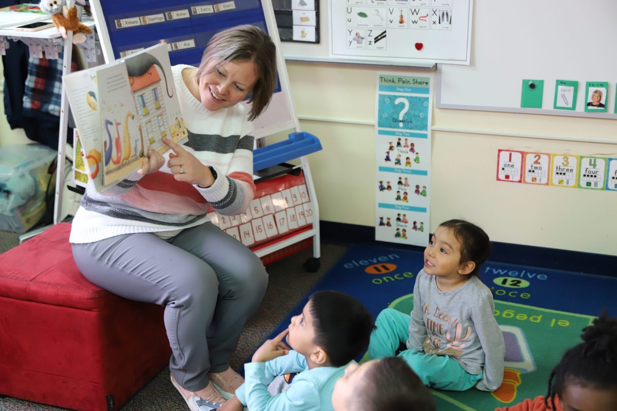 A teacher points to an illustration in a book as pre-schoolers seated around her look on
