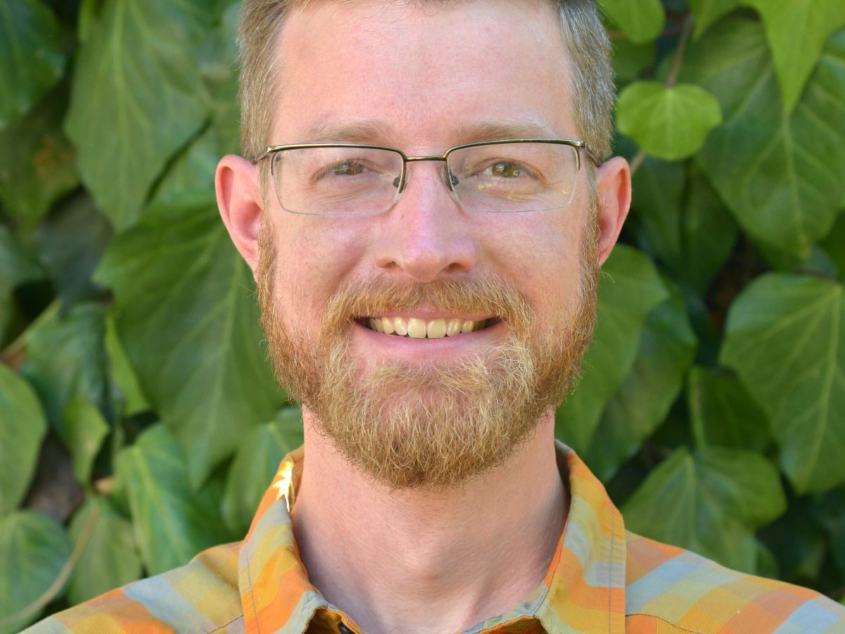 headshot of a smiling man standing in front of green vegetation