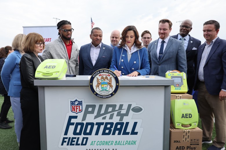 Gretchen Whitmer signs a bill while surrounded by a group of people and AEDs